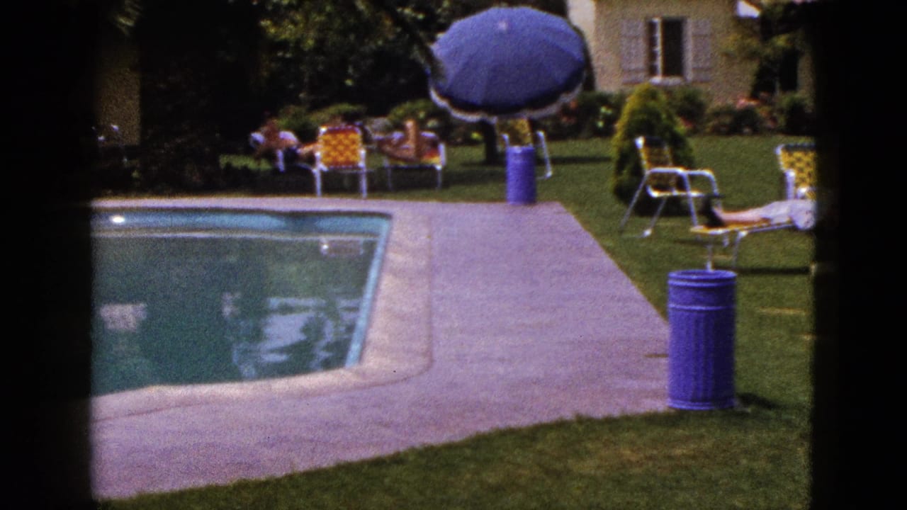 1953 New Mexico backyard pool scene with patio furniture, a large blue umbrella, green lawn, shrubs and a stucco house in the background.
