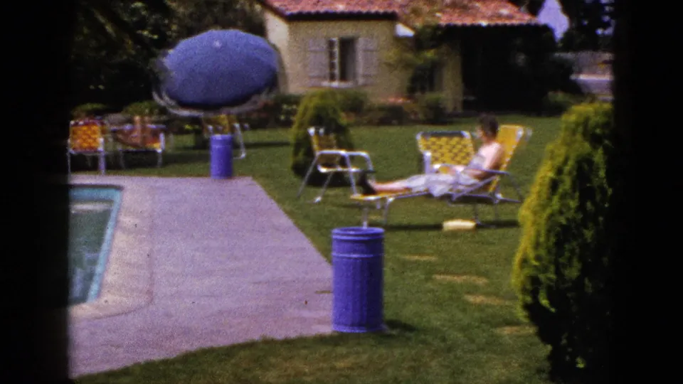 1953 New Mexico home-movie frame with a person reclining on a yellow lounge chair beside the lawn, pool deck, blue umbrella and shrubs.