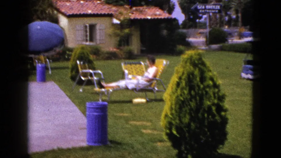1953 New Mexico 8mm frame showing a person seated on a yellow lounge chair near a pool patio, shrubs, umbrella and stucco house.
