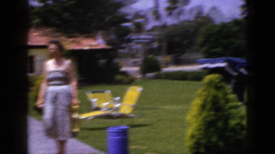 Soft-focused 1953 New Mexico frame of a woman moving along a concrete path beside a sunny motor hotel lawn.