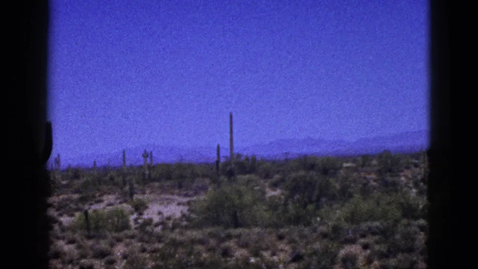 1953 New Mexico 8mm frame showing open desert terrain with scrub, small cacti, distant mountains, a blue sky, and dark border framing.