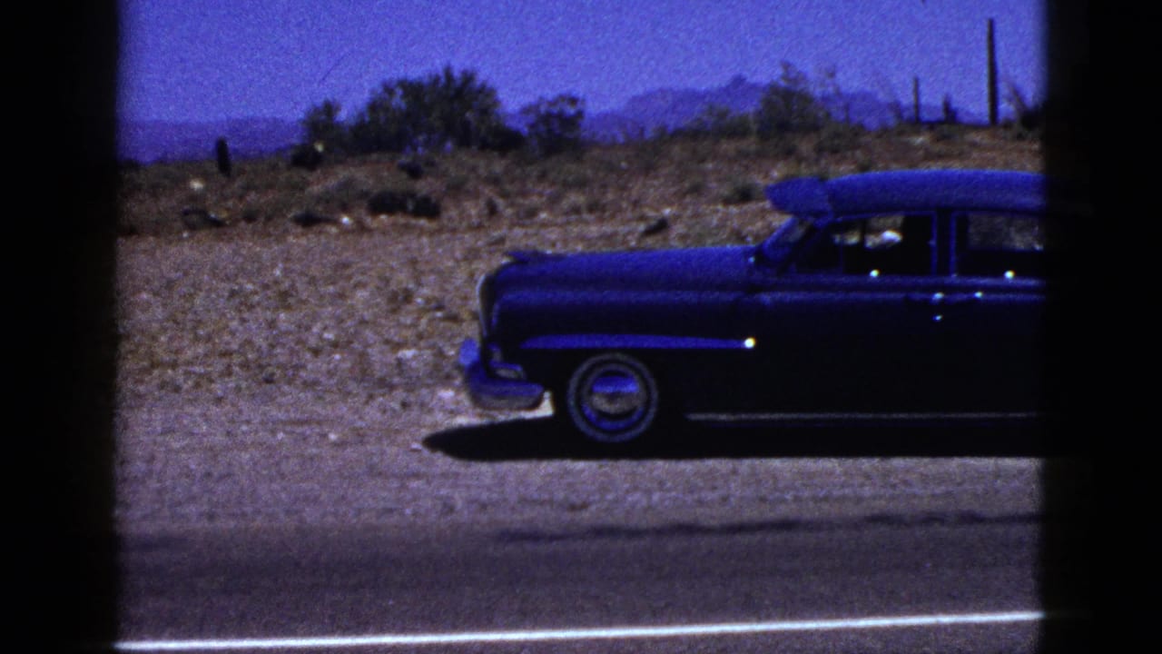 Front portion of a dark blue vintage sedan beside a desert road.