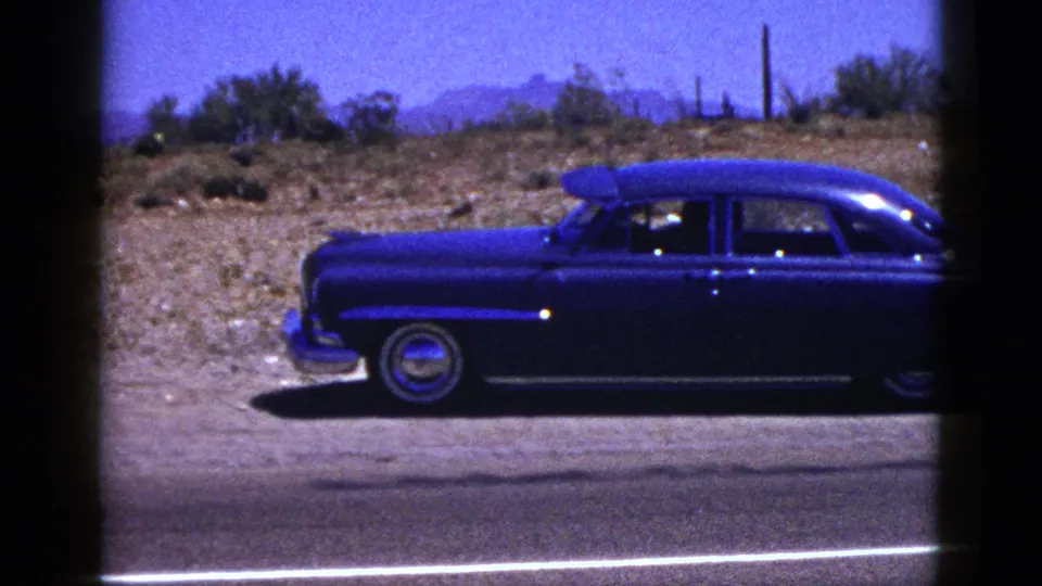 A dark blue mid-century sedan is shown in profile along a New Mexico roadside, with desert scrub and mountains in the background.