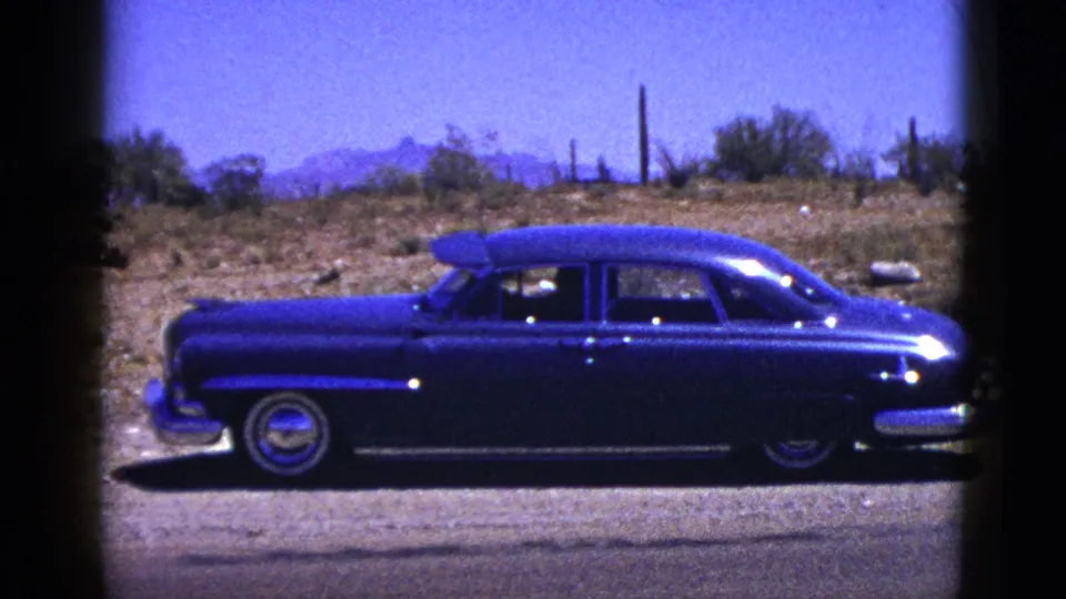 Fuller side view of the dark blue sedan parked on a New Mexico desert road, with scrub, cacti, and distant hills visible.