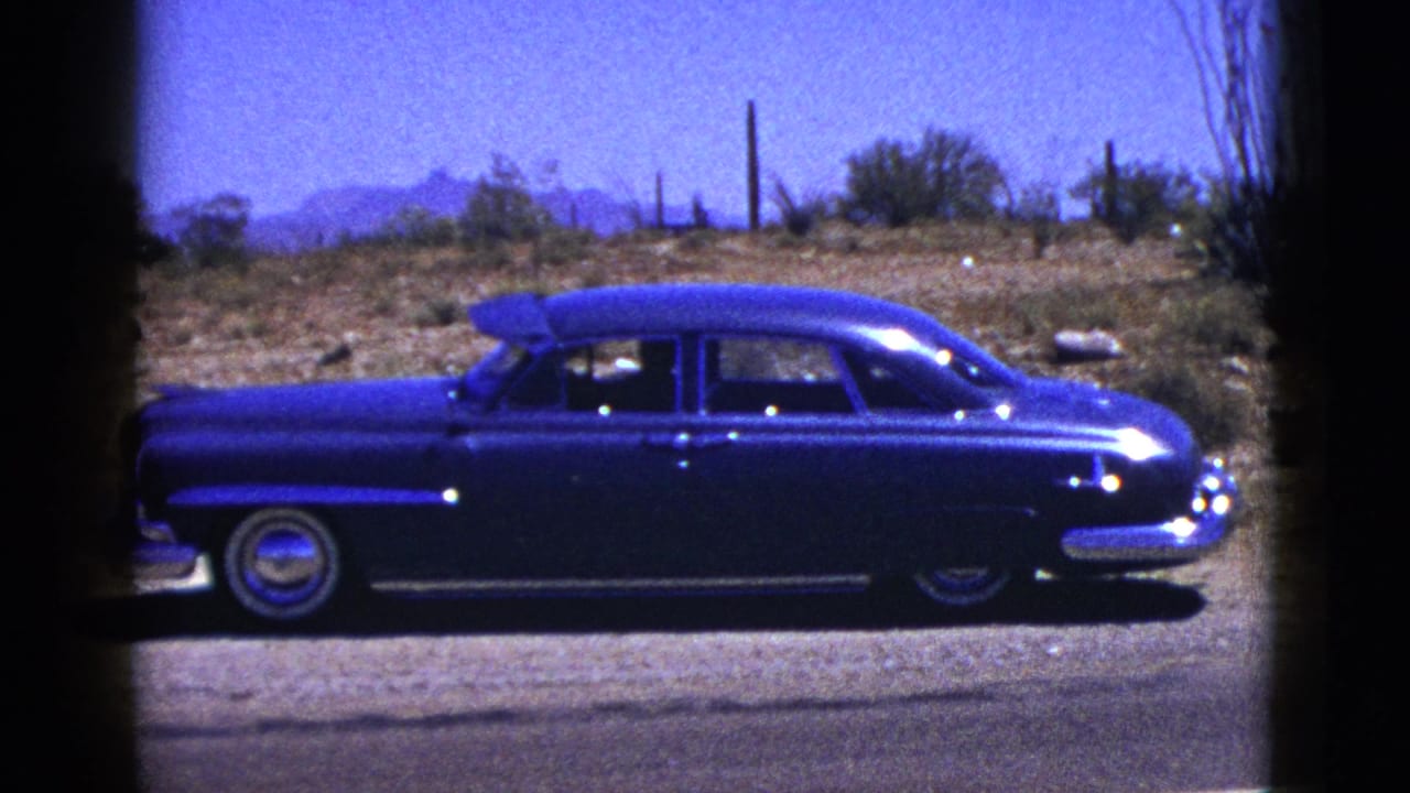 1953 New Mexico 8mm frame showing a blue mid-century sedan on a desert road shoulder with scrub, fence posts, and mountains beyond.