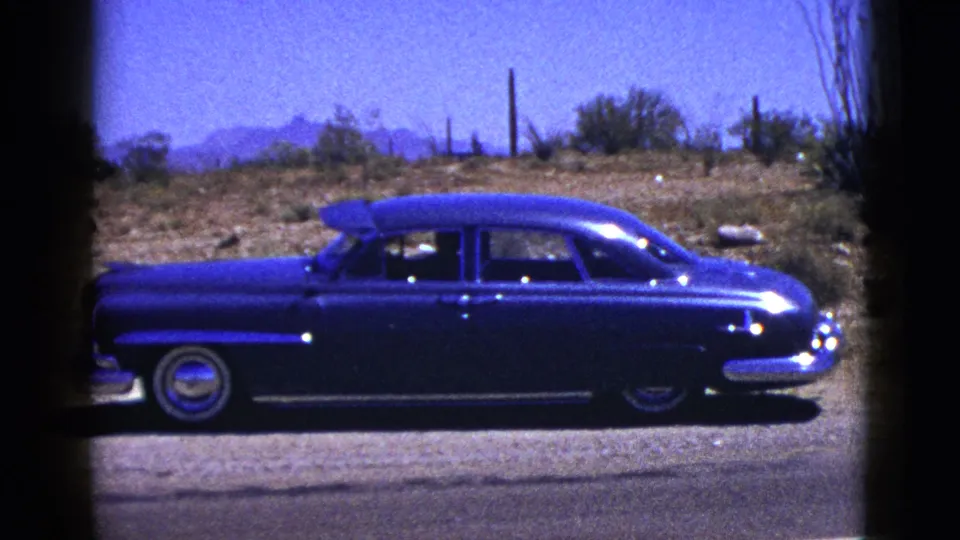 1953 New Mexico 8mm frame showing a blue mid-century sedan on a desert road shoulder with scrub, fence posts, and mountains beyond.