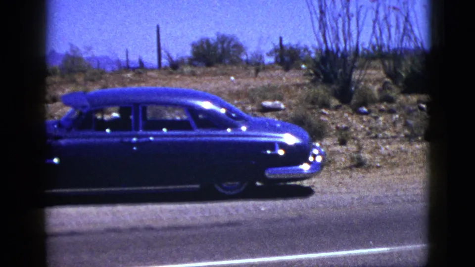 Side view of a blue 1950s sedan on a New Mexico desert road, with cacti or scrub, fence posts, and mountains in the background.