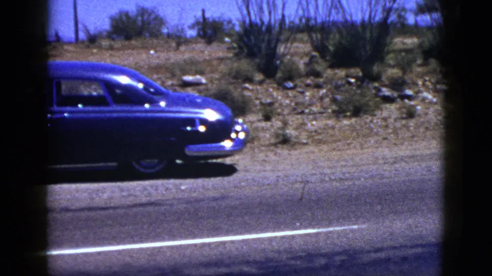 Partial 1953 New Mexico roadside view of the blue sedan, with pavement, desert scrub, rocks, and a bright sky visible through 8mm film edges.