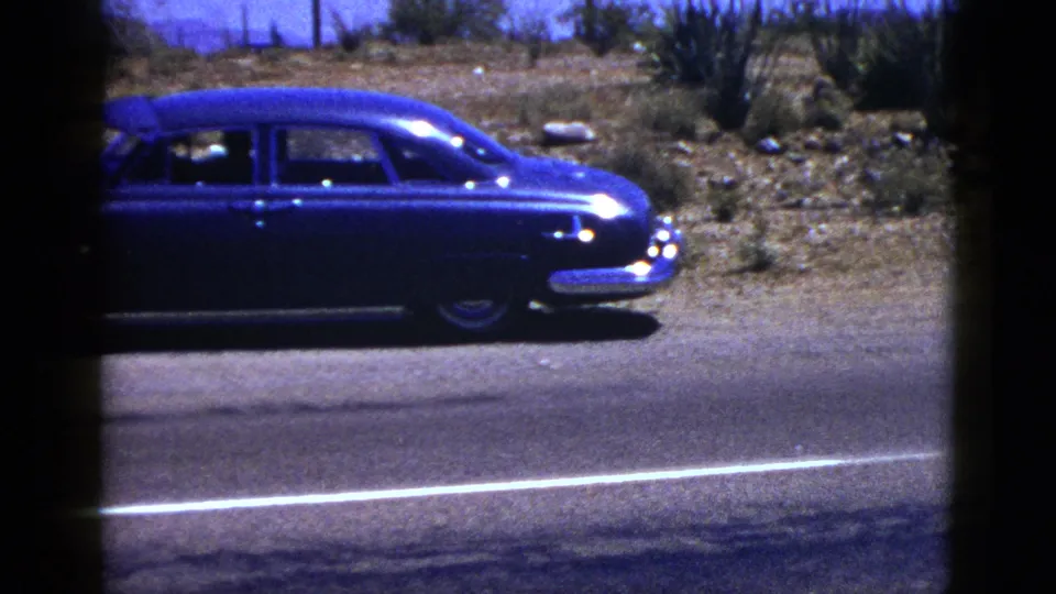Blue mid-century sedan on a New Mexico desert road shoulder in 1953, with chrome front detail, scrub plants, fence posts, and 8mm gate shadowing.
