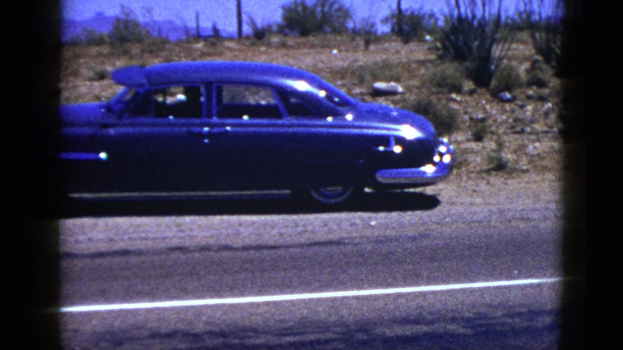 1953 Palm Desert 8mm frame showing a blue classic sedan moving along a desert highway under a clear sky.