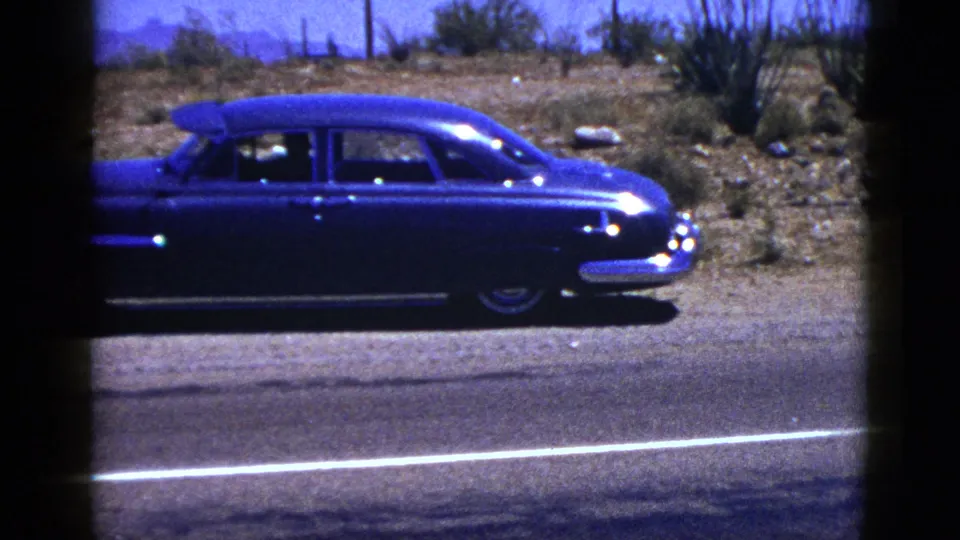 1953 Palm Desert 8mm frame showing a blue classic sedan moving along a desert highway under a clear sky.