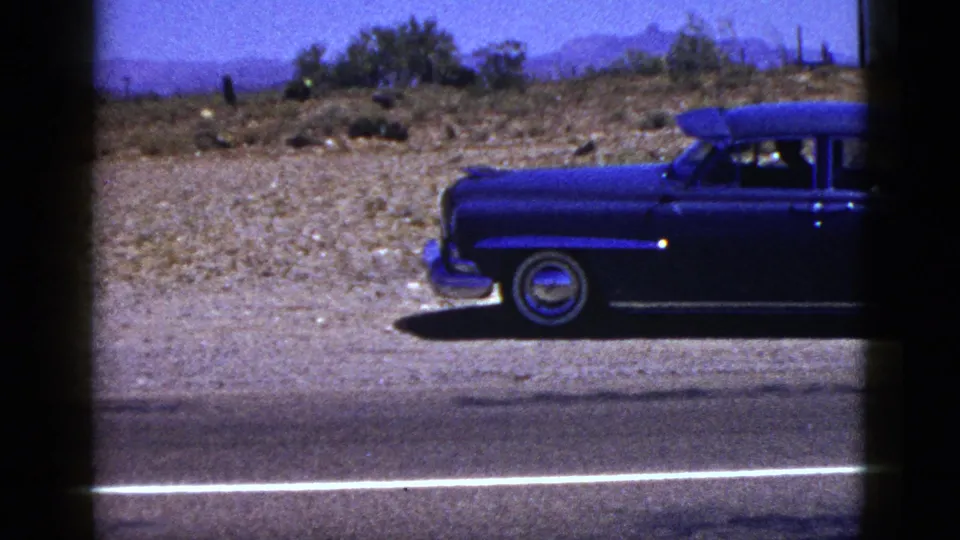 The blue sedan exits the 1953 Palm Desert highway frame, leaving dry scrubland and distant mountains behind.