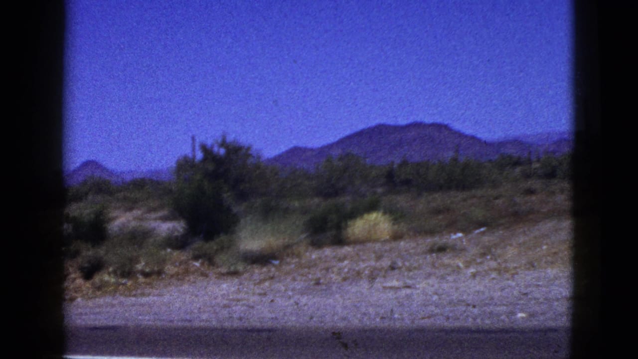 1953 Palm Desert desert terrain with scattered shrubs, rocky ground, and mountains beyond a roadside view.
