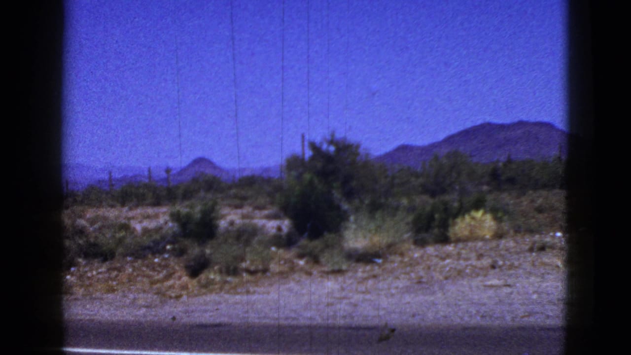 Roadside desert terrain with shrubs, mountains, and a dark vertical frame edge.