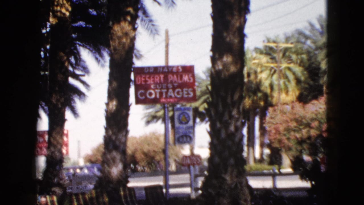 Red cottage sign, palms, road, and parked cars seen through tree trunks.