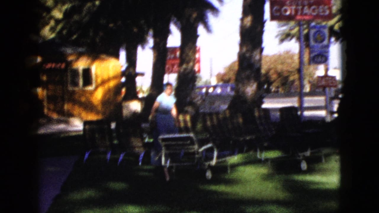 1953 Palm Desert motel grounds with a woman in blue near striped lounge chairs, palm trees, and red office and cottages signs.