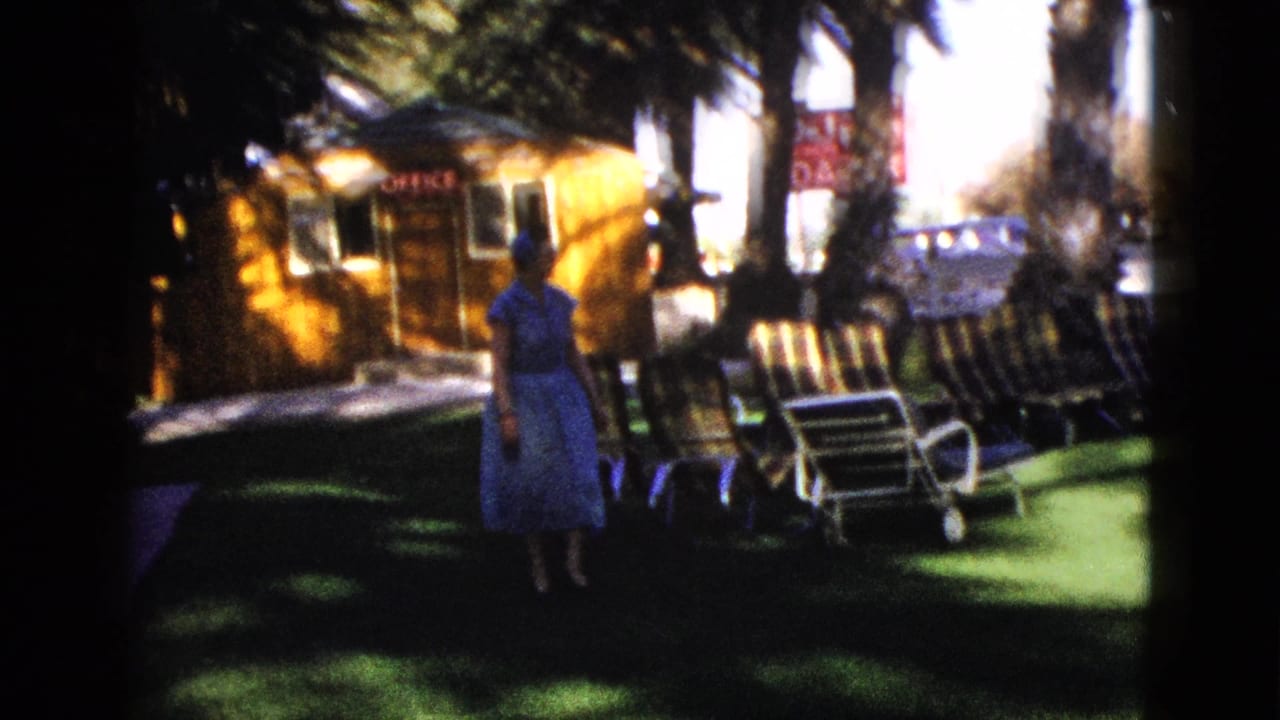 Woman stands on shaded lawn near lounge chairs with a small motel office building behind her.