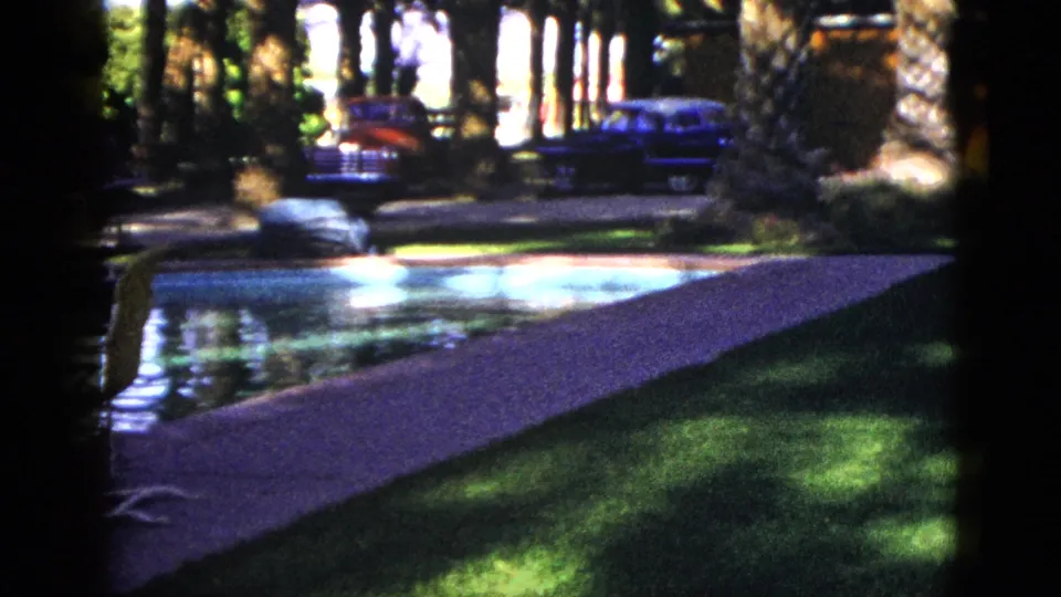 1953 Palm Desert motel grounds showing the pool edge, sunlit walkway, palms, lawn, and classic cars parked in the background.