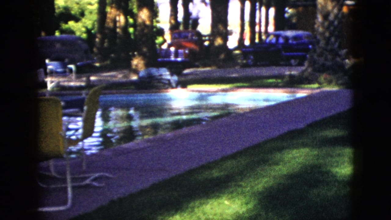 1953 Palm Desert home-movie frame showing a sunlit outdoor pool, green patio chair, trees, palms, and mid-century cars beyond the deck.