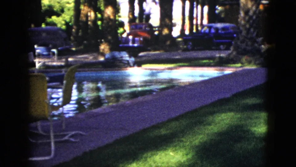 1953 Palm Desert home-movie frame showing a sunlit outdoor pool, green patio chair, trees, palms, and mid-century cars beyond the deck.