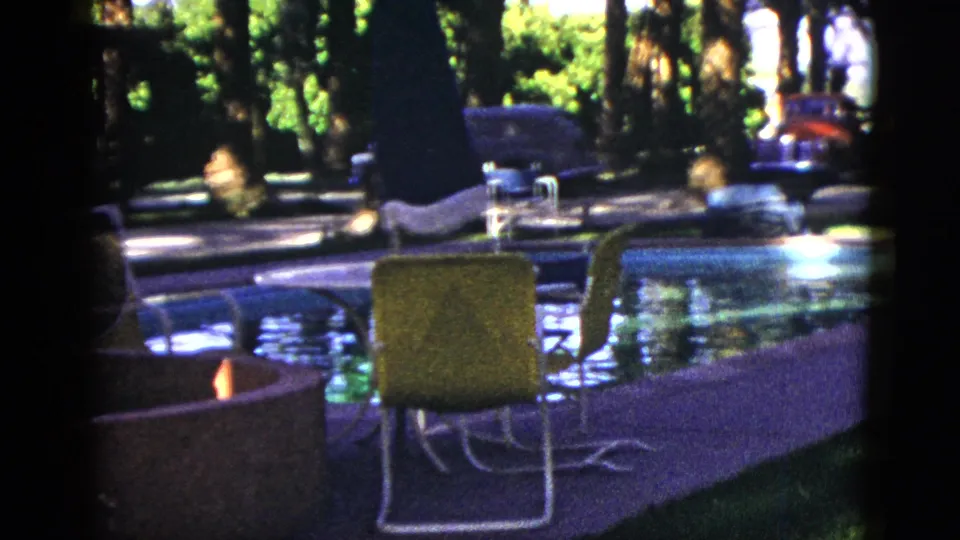 1953 Palm Desert frame showing patio furniture by the pool, a round fire pit or planter, shaded trees, palms, and parked cars.