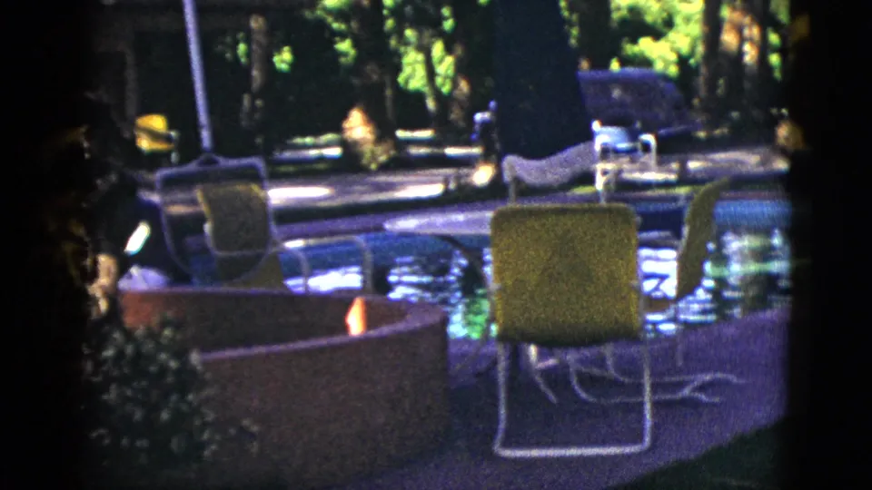 1953 Palm Desert pool scene with a green patio chair, round fire pit or planter, blue water, palm trunks, trees, and parked cars.