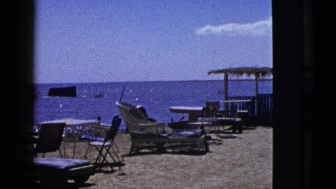 1953 archival 8mm view of a quiet California beach scene with wicker seating, folding chairs, tables, and calm blue water beyond.