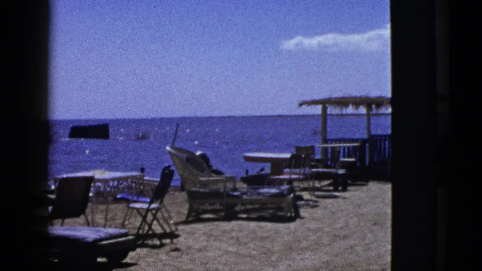 1953 archival 8mm view of a quiet California beach scene with wicker seating, folding chairs, tables, and calm blue water beyond.
