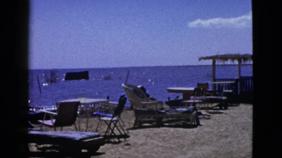 1953 California home-movie frame showing beach furniture arranged near calm water, with a thatched shade structure at the right edge.