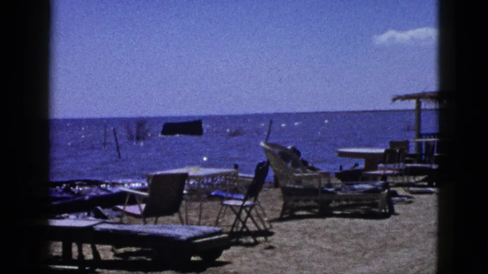1953 archival beach view with lounge furniture, folding chairs, scattered tables, and a dark floating object visible on the water.