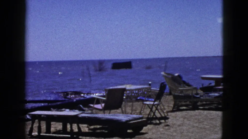 1953 California 8mm frame looking past a low lounge chair toward wicker seating, folding chairs, and a calm shoreline.