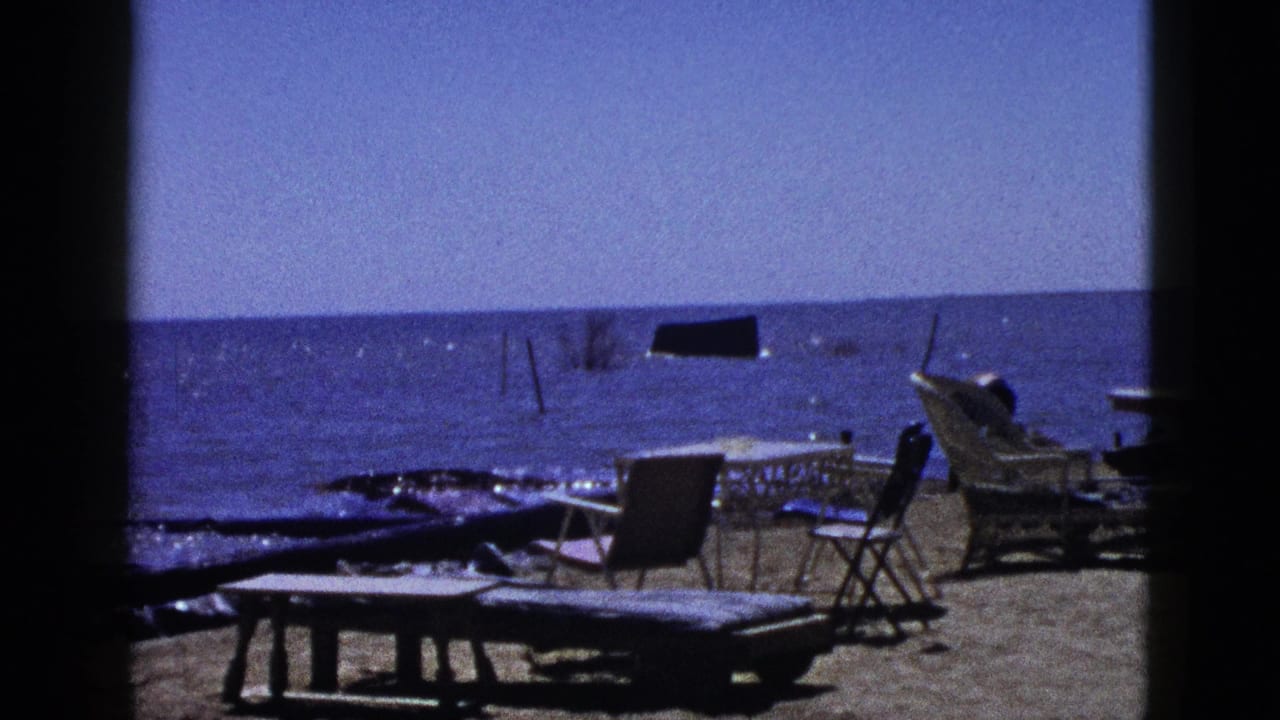 1953 California 8mm frame showing a sandy waterside area with driftwood, folding chairs, a low table, and bright blue water.