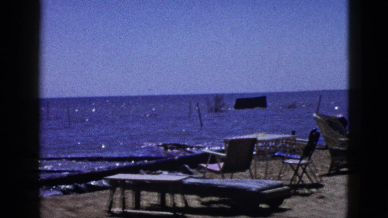 1953 California home-movie view of folding chairs, driftwood, and calm blue water seen through a dark foreground frame.