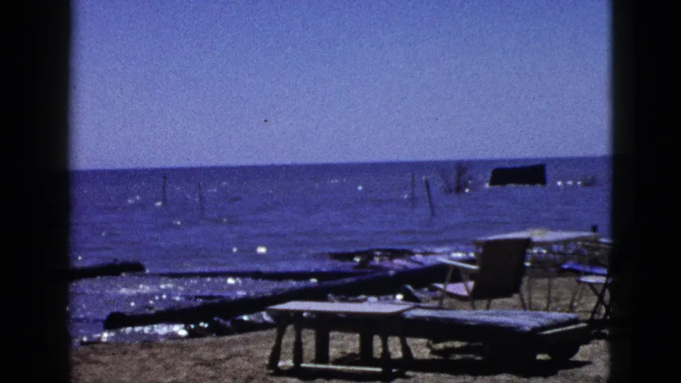 1953 California shoreline frame with a low table in front of driftwood, folding chairs, sand, and sparkling blue water.