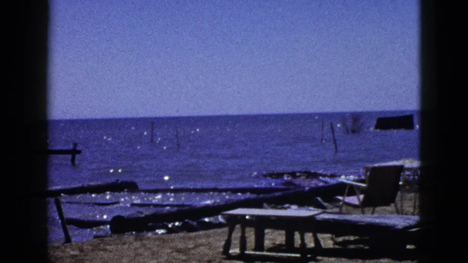 1953 California 8mm frame showing driftwood, a low table, chairs, and a calm sunlit shoreline through dark vertical framing.