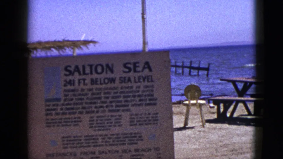1953 8mm frame showing the Salton Sea 241 ft. below sea level sign, picnic table, and shoreline water in Palm Desert metadata context.