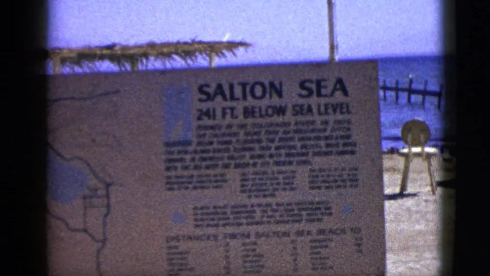 1953 8mm frame showing the Salton Sea 241 ft below sea level sign close to camera, with blue water and shoreline fixtures behind it.