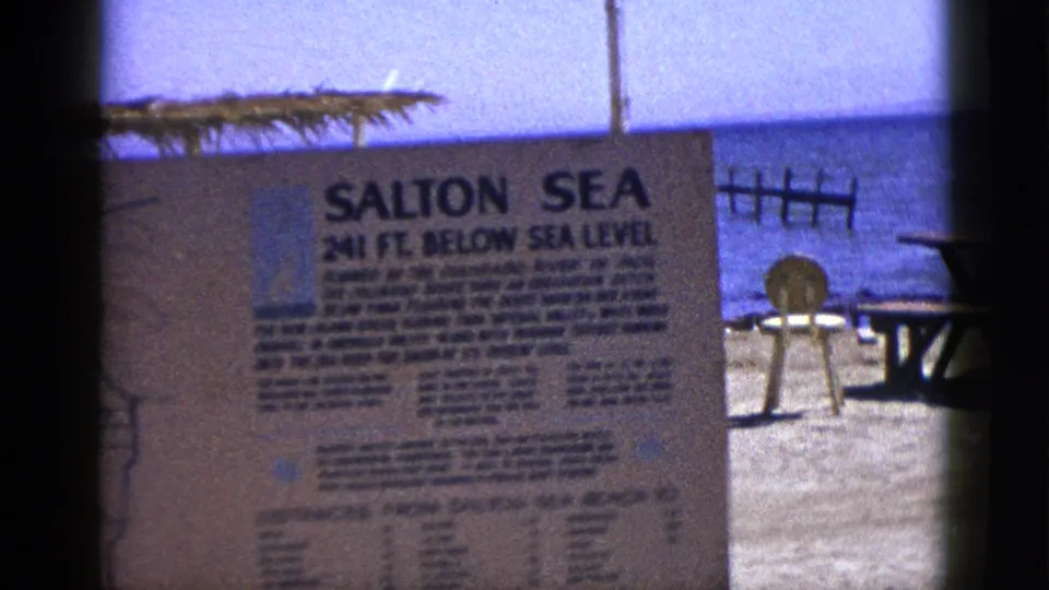 1953 8mm visitor-shot view of the Salton Sea sign, partially cropped, with shoreline, blue water, a round marker, and picnic table behind it.
