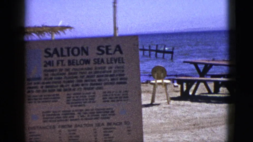1953 8mm frame of the Salton Sea sign on a sandy shore, with a picnic table, small round marker, posts, and calm blue water beyond.
