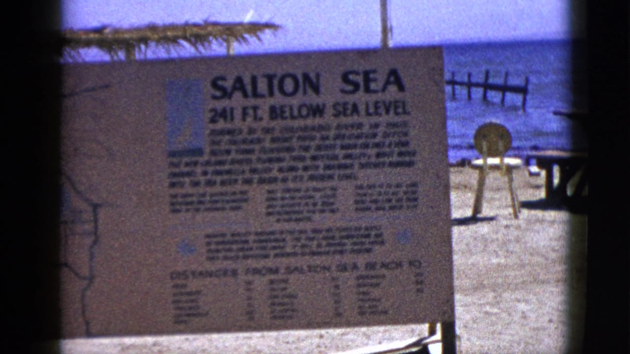 1953 8mm frame of the Salton Sea beach sign, with the shoreline, a small chair, and a wooden structure visible under bright daylight.