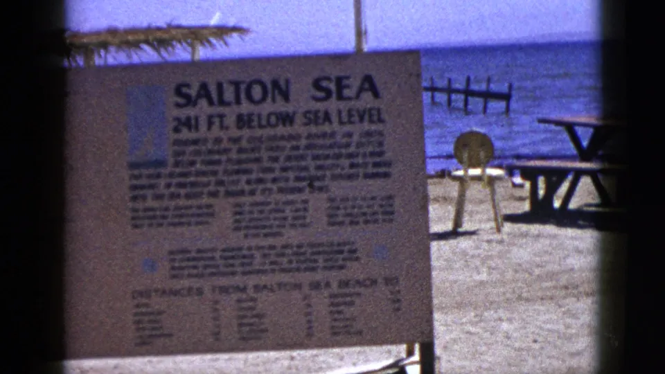 1953 8mm frame showing the Salton Sea informational sign in front of beach sand, with a picnic table, chair, and blue water visible to the side.