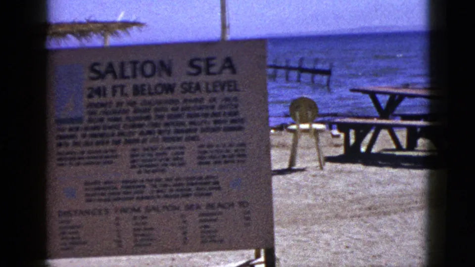 1953 8mm frame of the Salton Sea sign on a sandy beach, with a picnic table, chair, wooden shoreline structure, and blue water behind it.