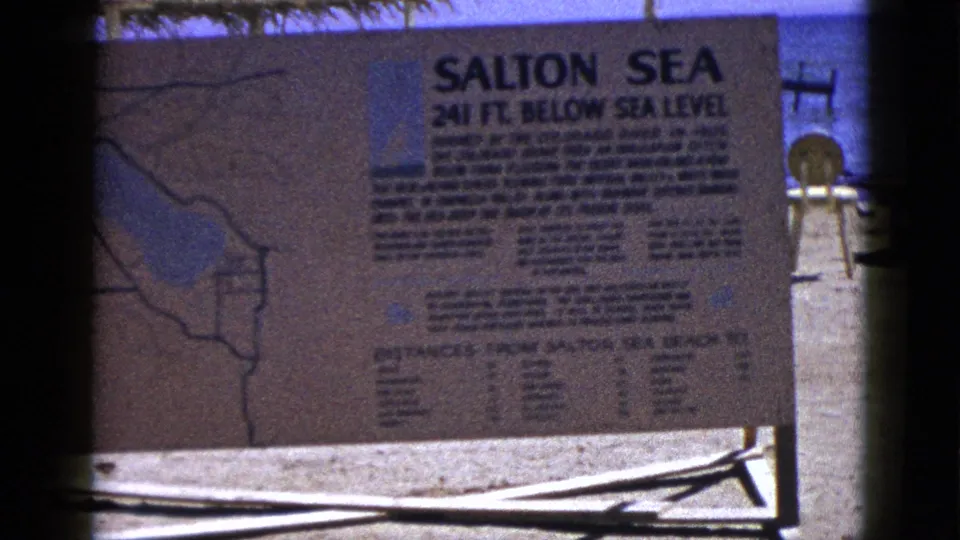 1953 8mm view of a Salton Sea sign noting 241 feet below sea level, with water and shore structures behind it.