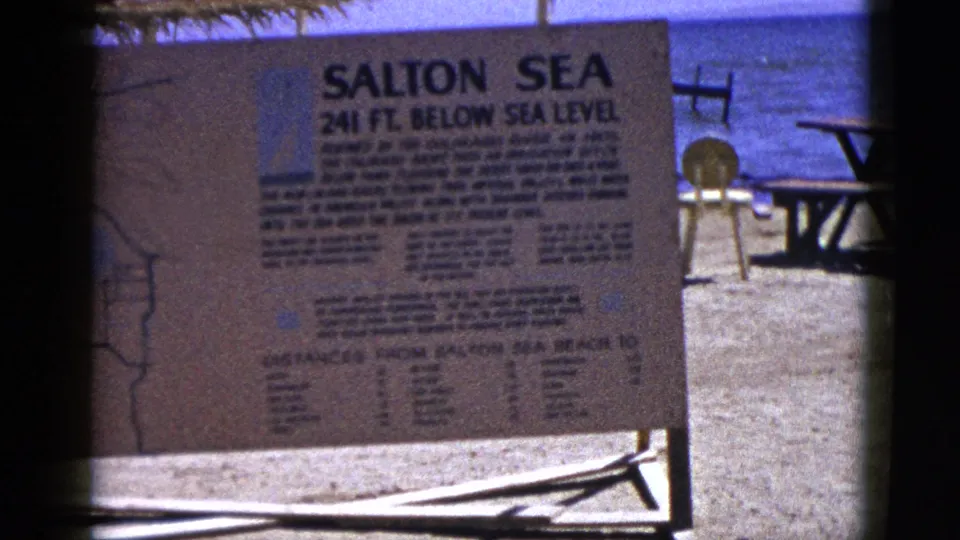 1953 Palm Desert-area home-movie frame showing the Salton Sea sign, below-sea-level text, and calm water beyond the shore.