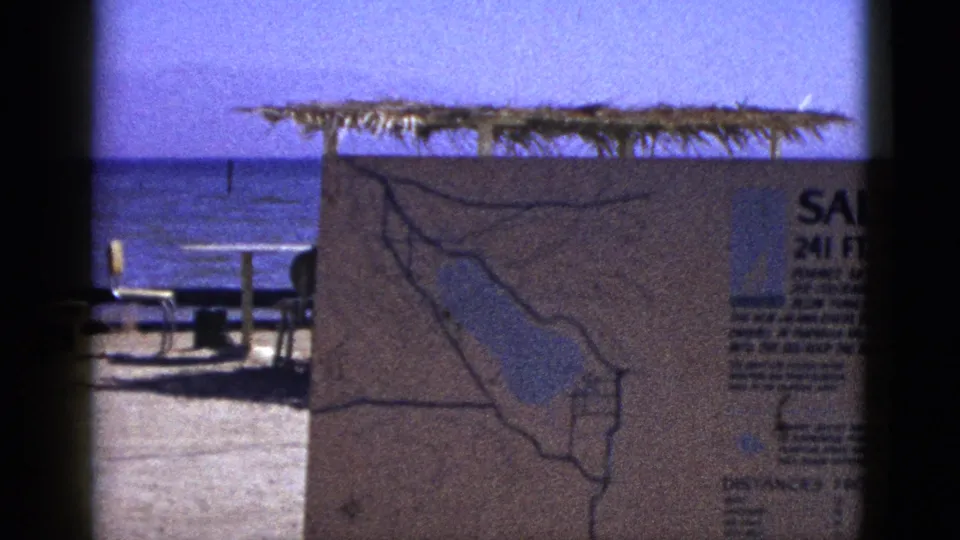 1953 8mm frame emphasizing the sign's regional map, with picnic tables, sandy ground, and the Salton Sea in the background.