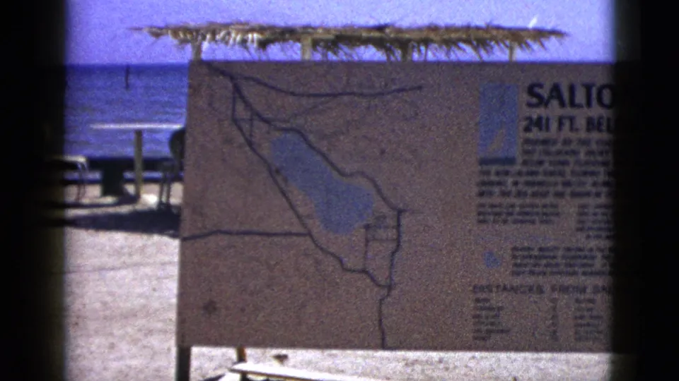 1953 Palm Desert-area frame showing the map side of the Salton Sea sign, with thatched shade, picnic tables, and water behind it.