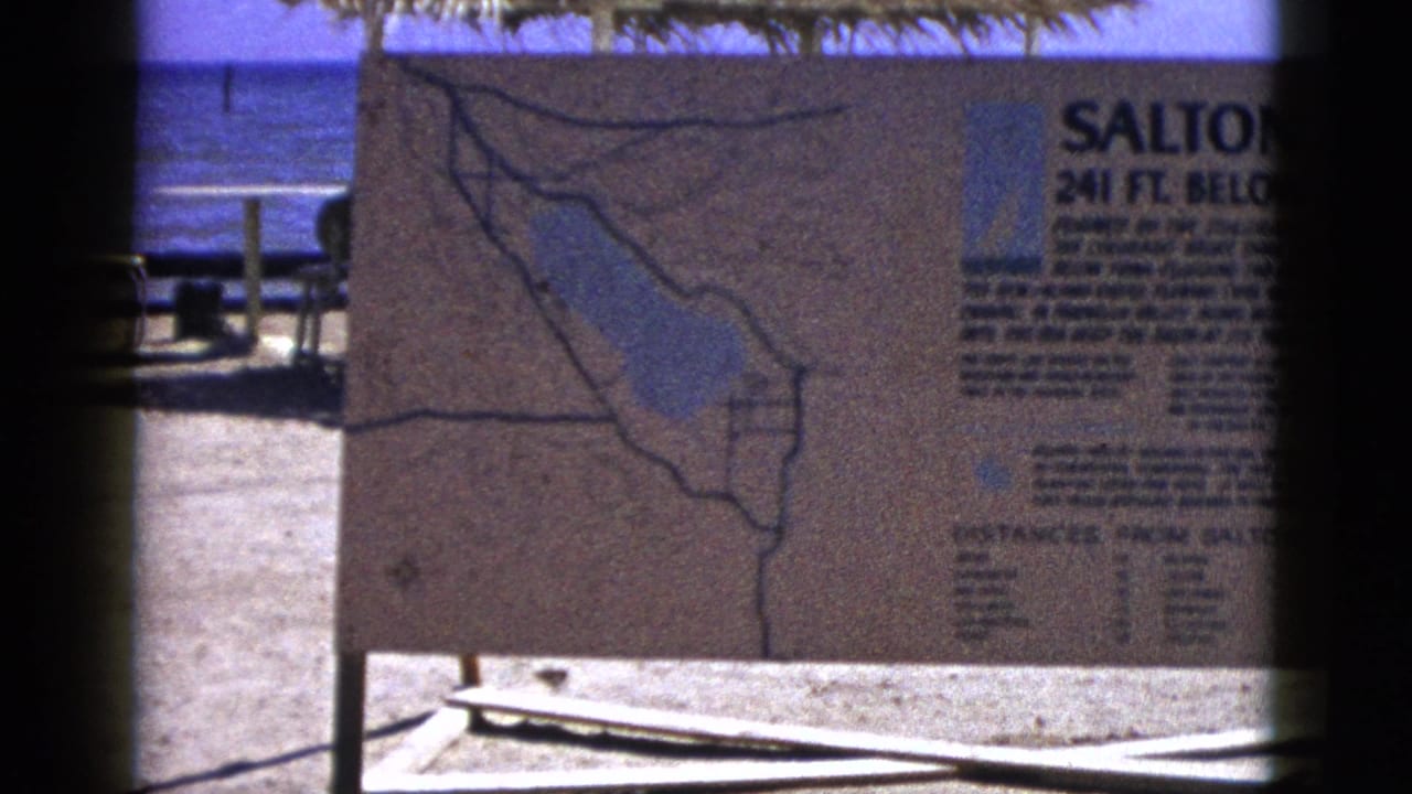 Salton Sea informational sign with map beside blue water and sandy ground.