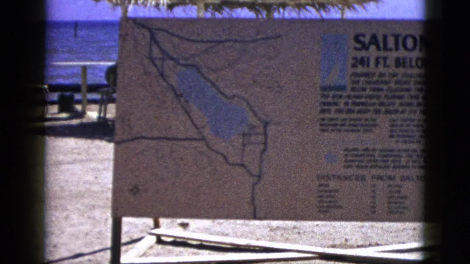 1953 8mm frame of a Salton Sea informational sign with a map, sandy foreground, shade roof, and blue water behind it.