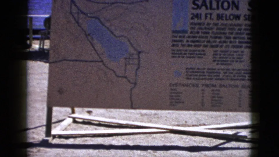 1953 8mm view of the Salton Sea display sign showing a map outline, text blocks, posts, and distant blue water.
