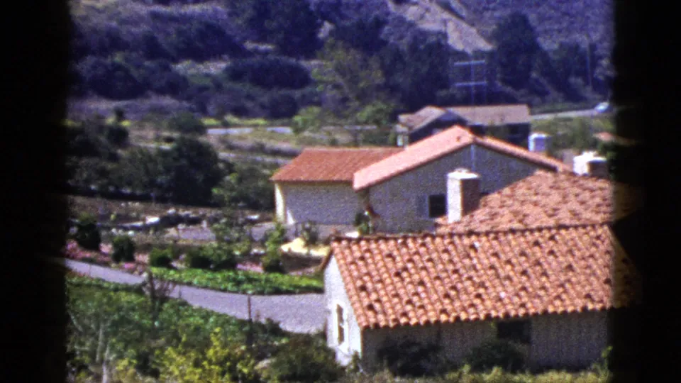 1953 8mm frame of Palm Springs area homes with red tile roofs, pale walls, landscaping, and hills in the background.